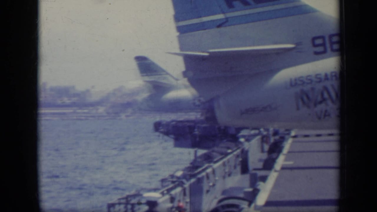 Carrier deck beside open water with parked aircraft tail marked USS Saratoga and Navy text.