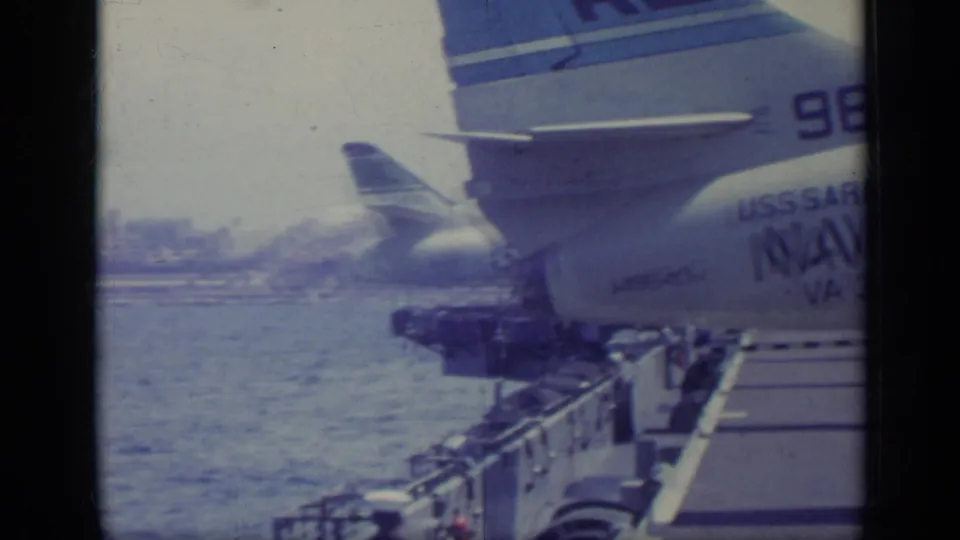1962 Italy 8mm frame showing a U.S. Navy carrier deck, water, railings, and a parked aircraft tail with USS Saratoga markings.