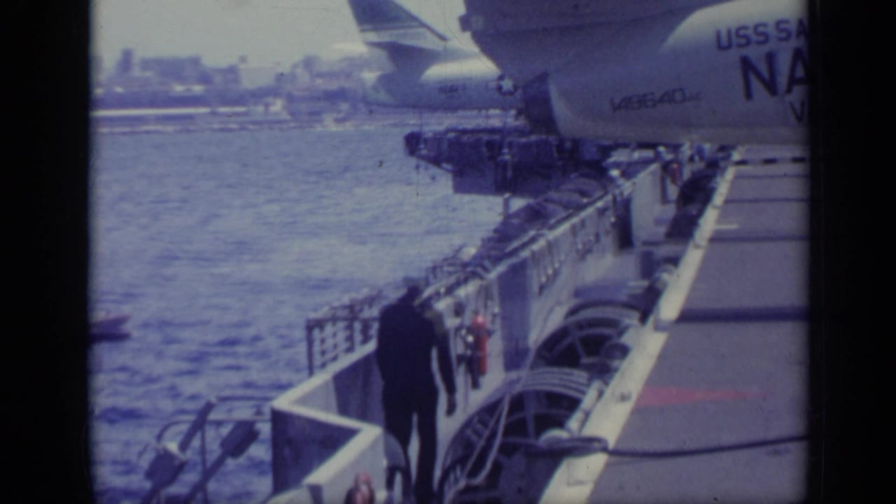 Sailor walks along the side of a carrier deck near railings and parked aircraft.