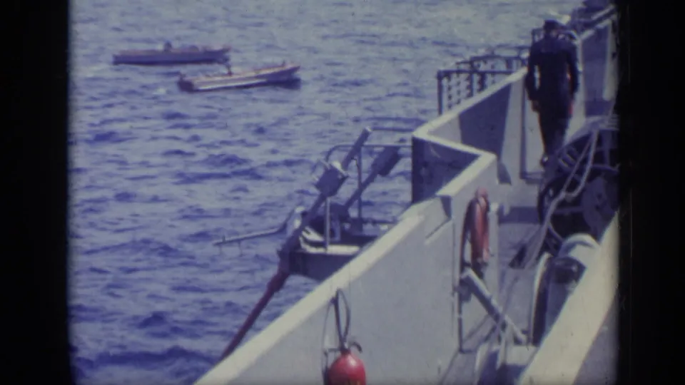 Handheld 8mm view from a carrier deck with a sailor near the railing, small boats on the water, and ship equipment in view.