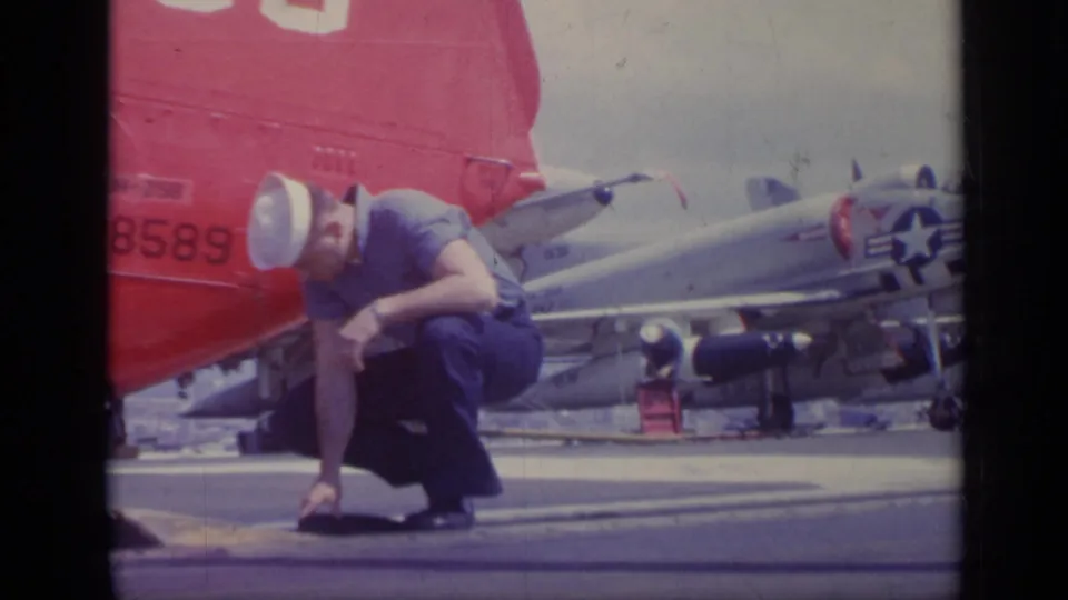 A sailor in a white cap and blue uniform crouches near aircraft on the USS Saratoga flight deck, with naval markings visible.