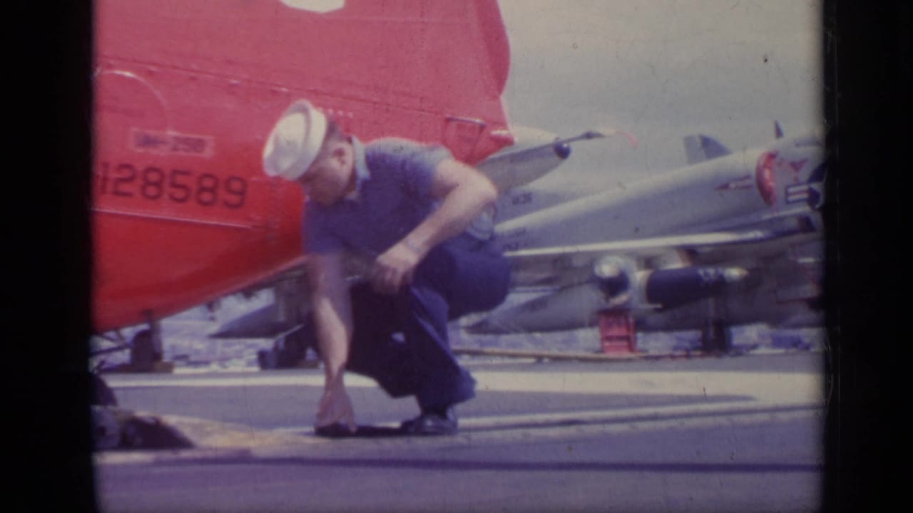 U.S. Navy sailor crouching beside a red helicopter on an aircraft carrier deck.