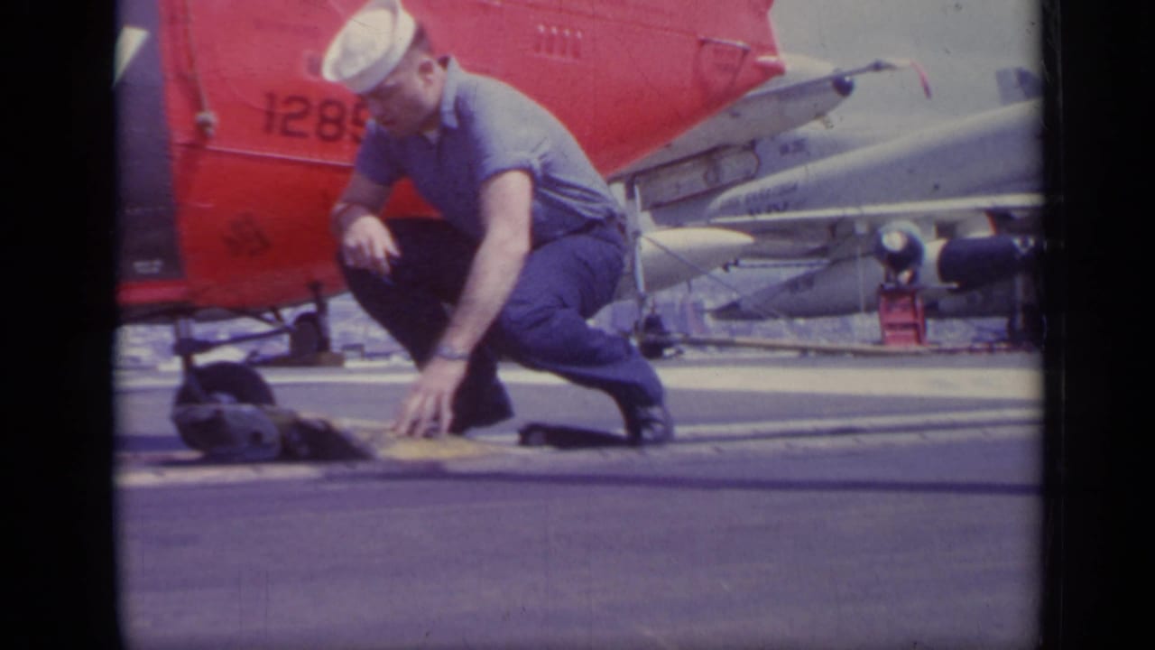 A U.S. Navy sailor crouches on the carrier deck near small equipment, with a red helicopter tail and jet aircraft visible behind him.