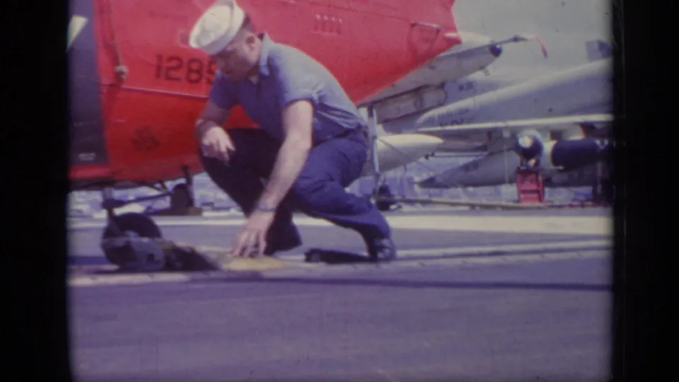 A U.S. Navy sailor crouches on the carrier deck near small equipment, with a red helicopter tail and jet aircraft visible behind him.