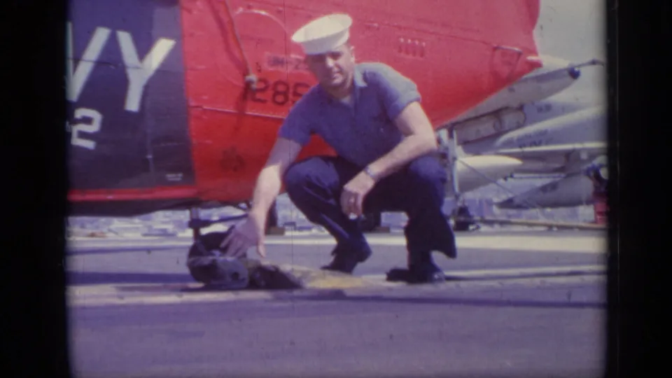 1962 Italy home-movie frame of a sailor crouched and facing the camera beside a red helicopter tail marked with numbers.