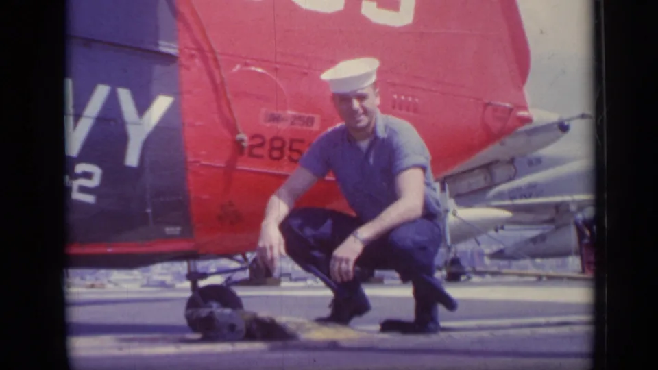 A U.S. Navy sailor poses in a crouch beside deck equipment, with a red helicopter tail and nearby aircraft in the background.