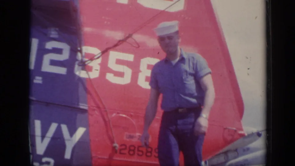 In Italy, 1962, a sailor stands on the deck in front of the helicopter tail with large white numbers and letters visible.