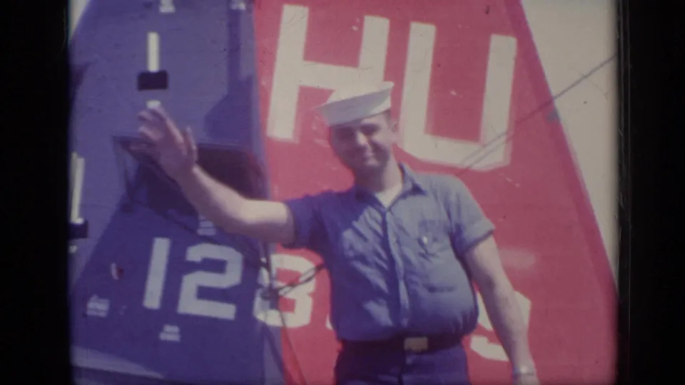 In Italy, 1962, a sailor raises one hand in a wave while standing beside the red and blue Navy helicopter tail.