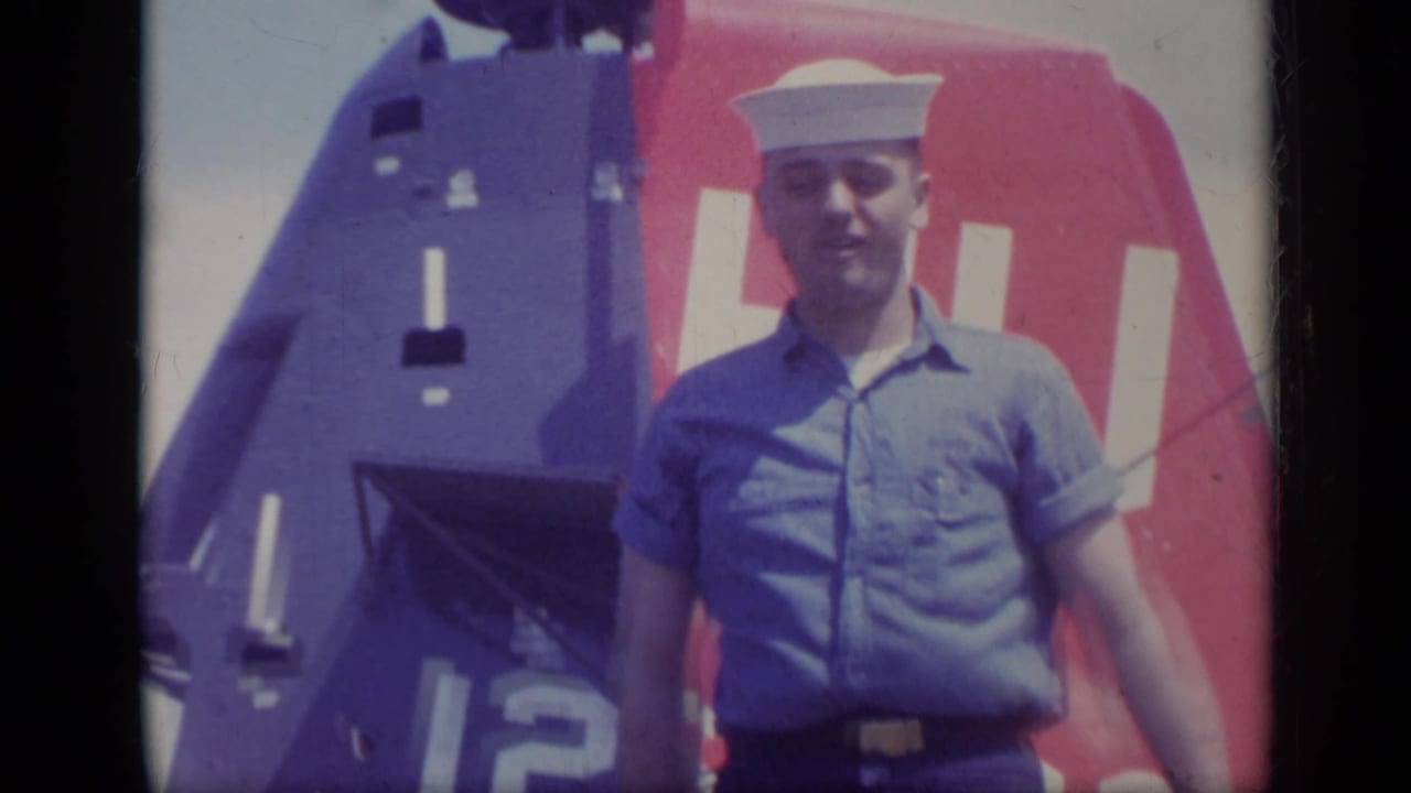 A U.S. Navy sailor poses in uniform before a marked helicopter on a carrier deck in metadata-listed Italy, 1962.