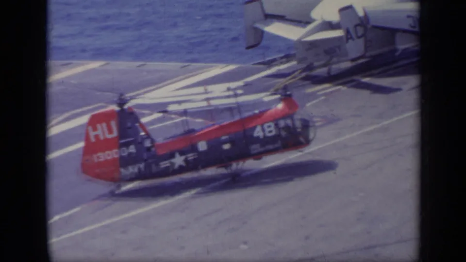 A red and blue Navy helicopter sits on the aircraft carrier deck, with painted deck lines and the sea in the background.