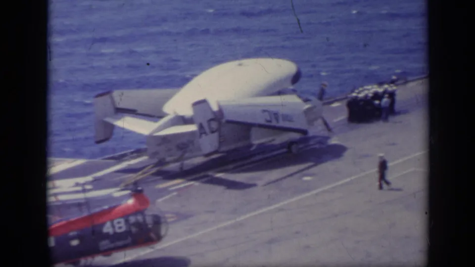 Wide 1962 carrier-deck view shows a helicopter, radar-equipped aircraft, small crew figure, and open water beyond.