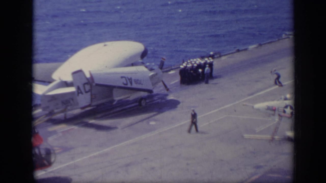 Carrier flight deck with a propeller aircraft, sailors, parked jets, and ocean beyond the rail.