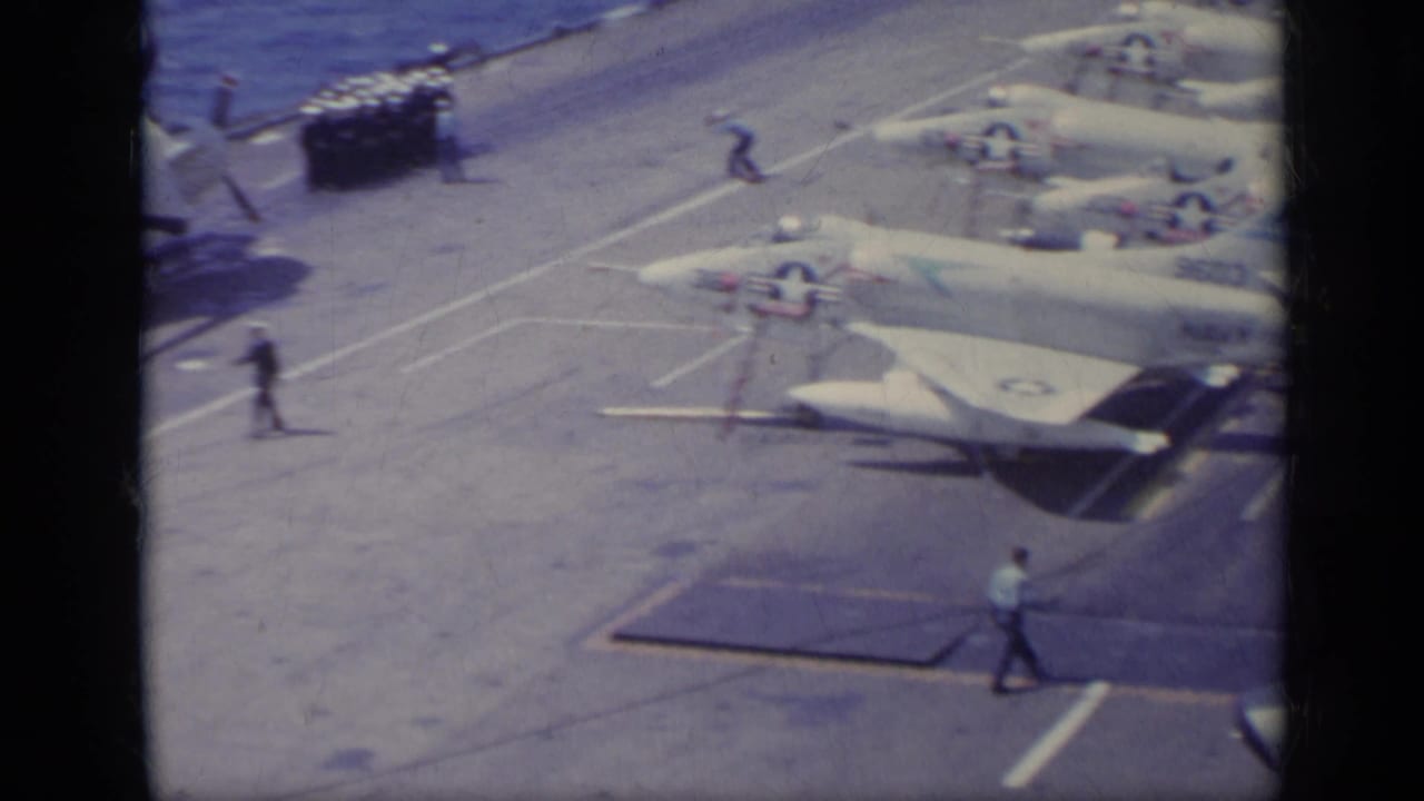 Parked Navy jets on a carrier deck with sailors walking and the ocean along the deck edge.
