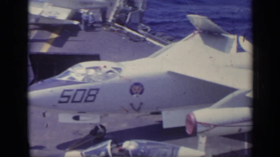 1962 Italy metadata-supported 8mm frame with a close U.S. Navy jet nose marked 508, cockpit canopy, deck edge, sailors, and ocean beyond.