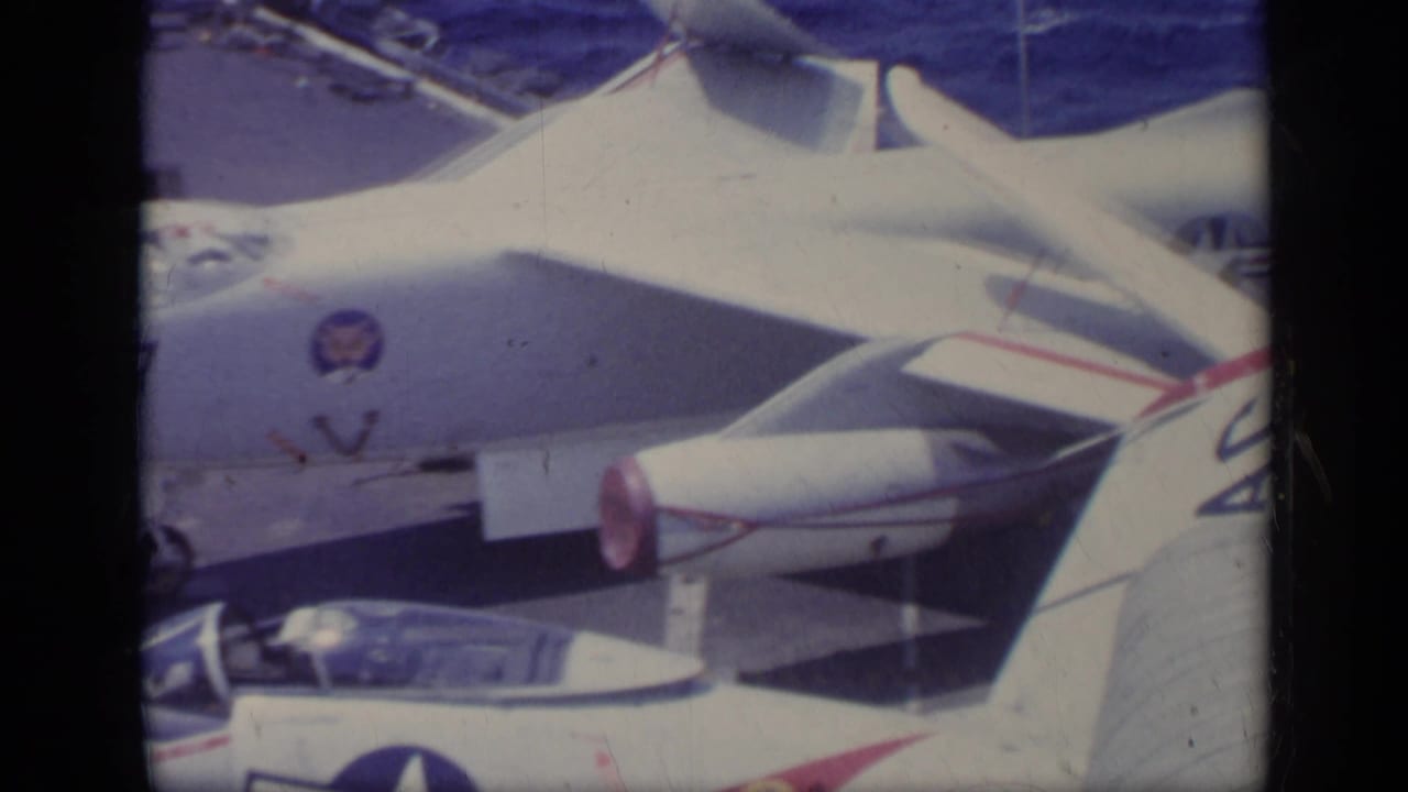 Close view of parked white naval jets on a carrier deck with the sea in the background.