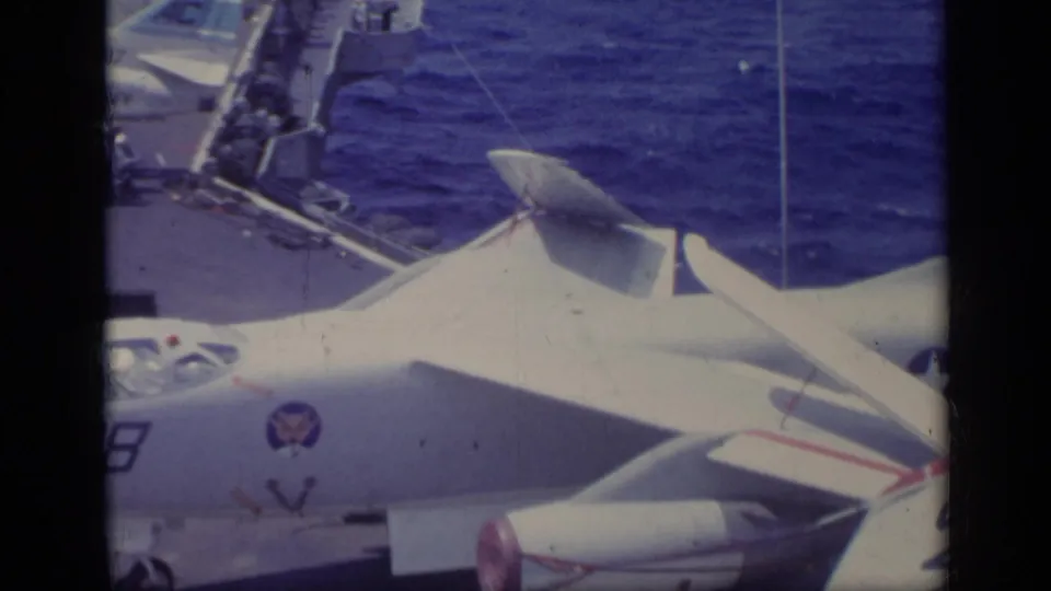 Overhead 8mm view of a white U.S. Navy jet on a carrier deck, with another aircraft edge and ocean visible behind it.