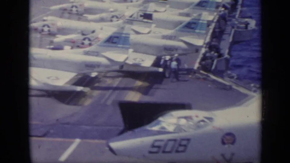 Wider 1962 Italy-associated carrier-deck frame with multiple white U.S. Navy jets, deck markings, ocean, and small visible crew figures.