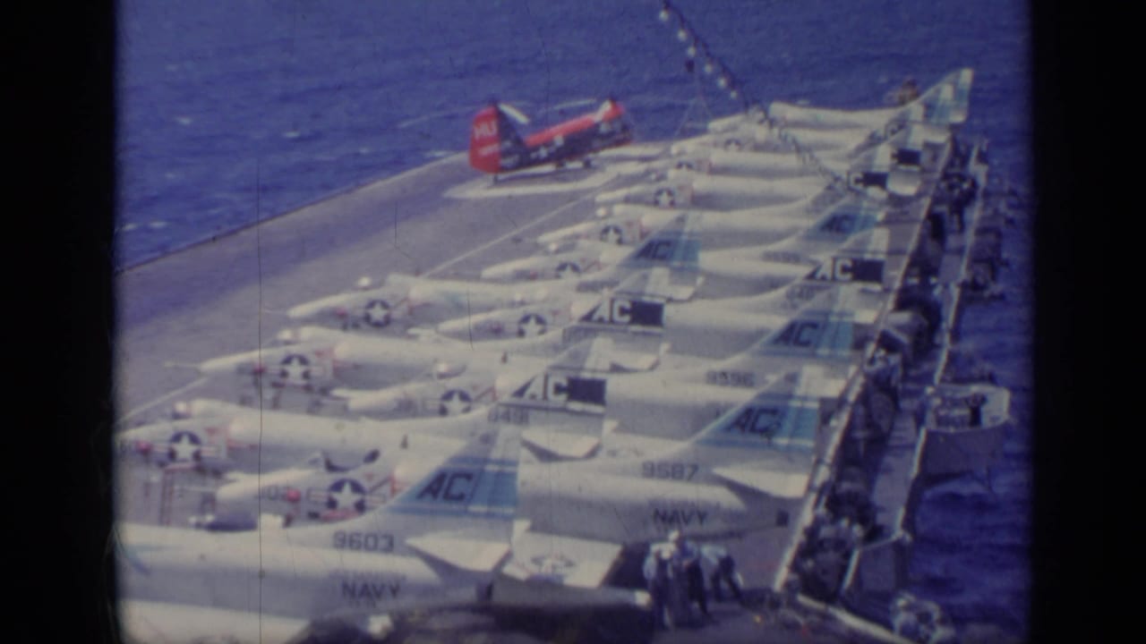 Carrier deck at sea with rows of jets and a red helicopter near the bow.