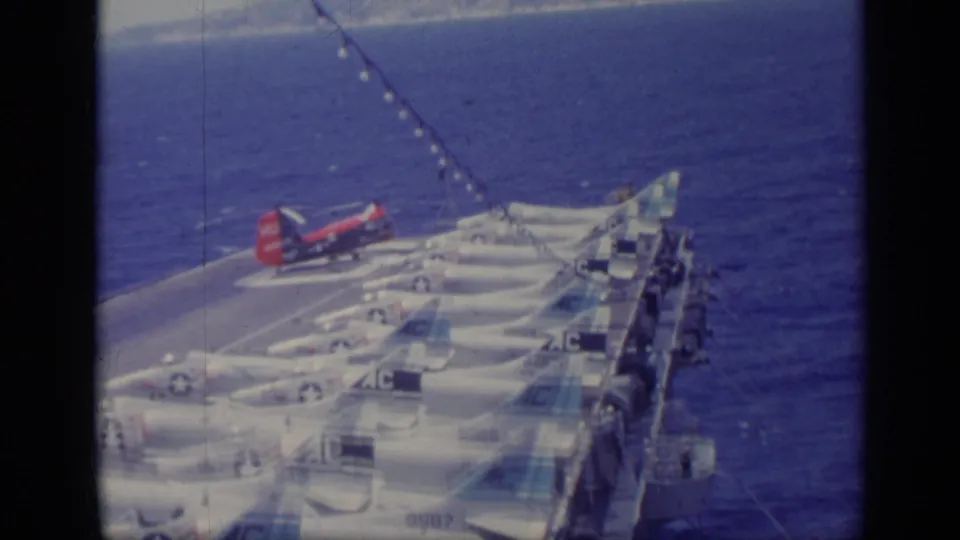 1962 Italy 8mm frame showing multiple Navy jets on a carrier deck with a red helicopter and sea beyond.