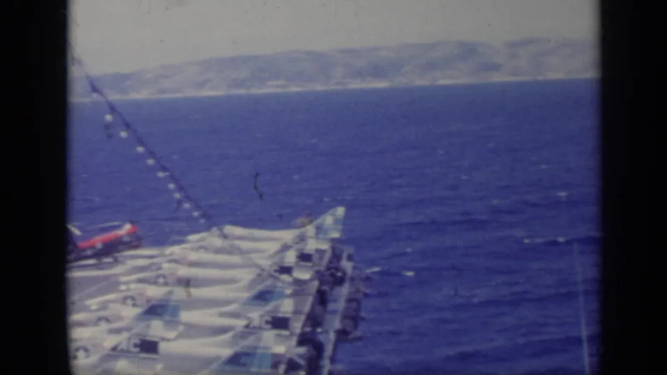 1962 Italy 8mm frame showing the forward carrier deck with parked jets, open water, and land on the horizon.