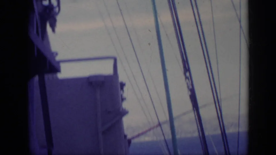 1962 Italy 8mm frame looking past ship structure and rigging toward blue water and a faint coastline.