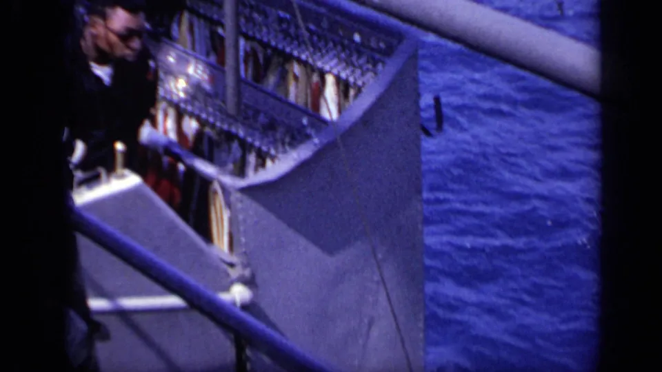 A partly visible crew member stands by freighter railings and deck fittings, with the blue ocean filling the background.