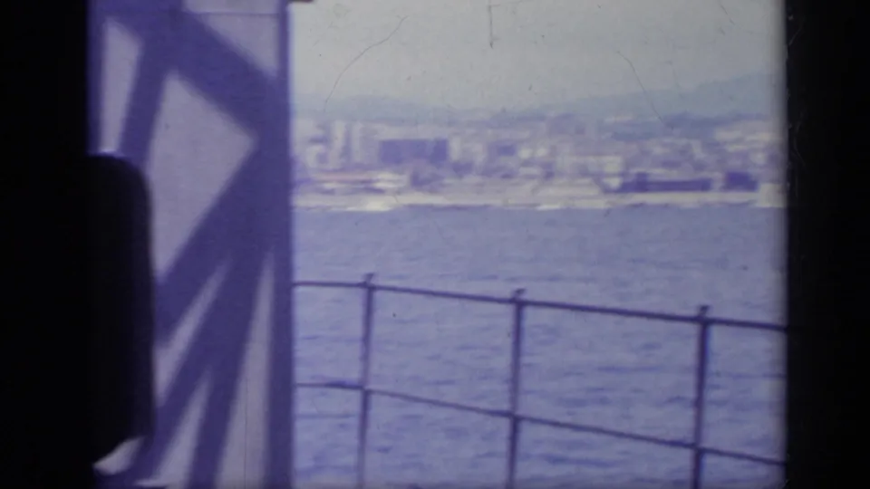 1962 Italy 8mm frame looking past a ship railing across water toward a hazy coastal city skyline.
