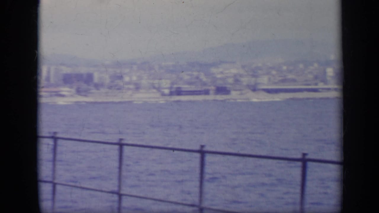 Blue water with a distant coastal city and mountains, seen past a vessel railing and dark frame edge.