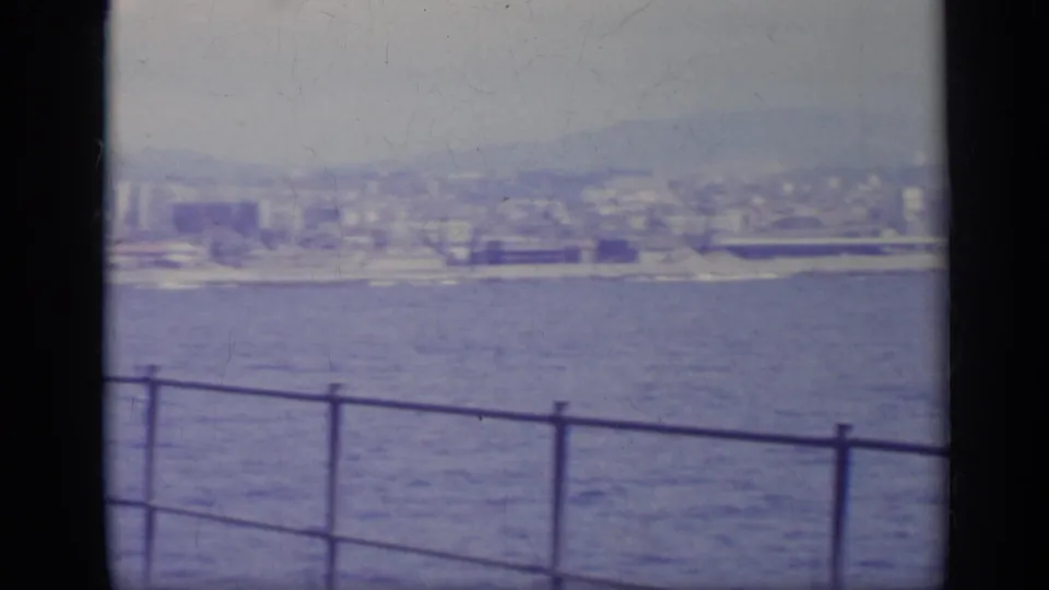 1962 Italy 8mm view across blue water toward a coastal city and mountains, with a metal railing and dark window edge in front.
