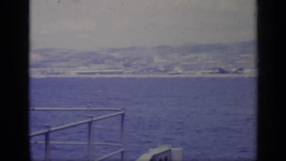 1962 Italy home-movie frame showing blue water, shoreline buildings, distant mountains, and a vessel railing beside a dark frame edge.