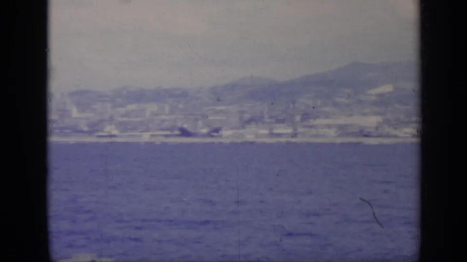 1962 Italy 8mm frame with a hazy coastal city and mountain ridge beyond blue water, bordered by dark film or window edges.