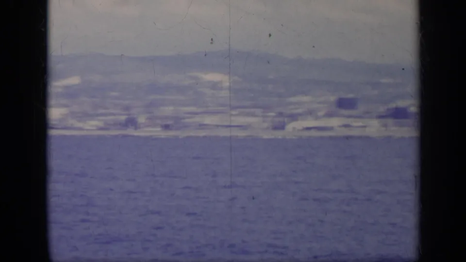 1962 Italy 8mm frame showing a distant coastal city and low hills viewed across open blue water.