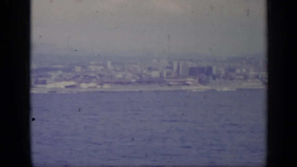 1962 Italy home-movie frame with a broad sea foreground and a distant urban waterfront under gray light.