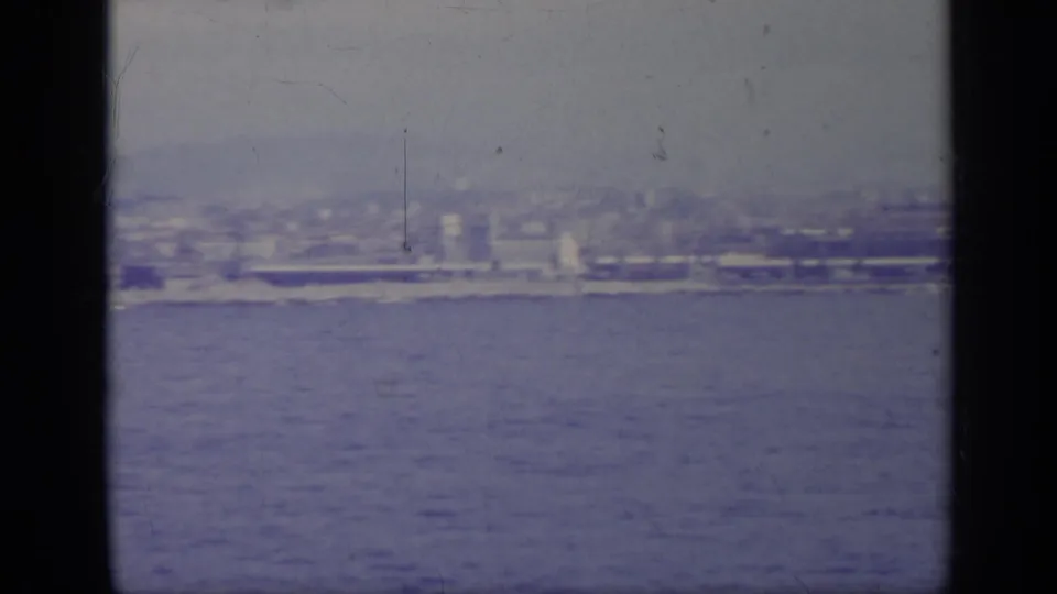 1962 8mm view of an Italian coastal city across calm water, partially blocked by dark vertical bars and softened by haze.