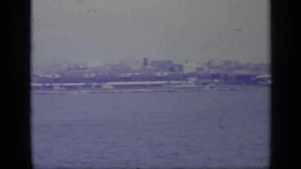 Distant Italian waterfront buildings and shoreline structures viewed across the sea on faded 1962 8mm film.