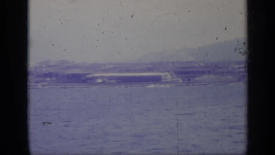 Soft 8mm frame of an Italian coastal shoreline across water, with low buildings and haze behind dark foreground bars.