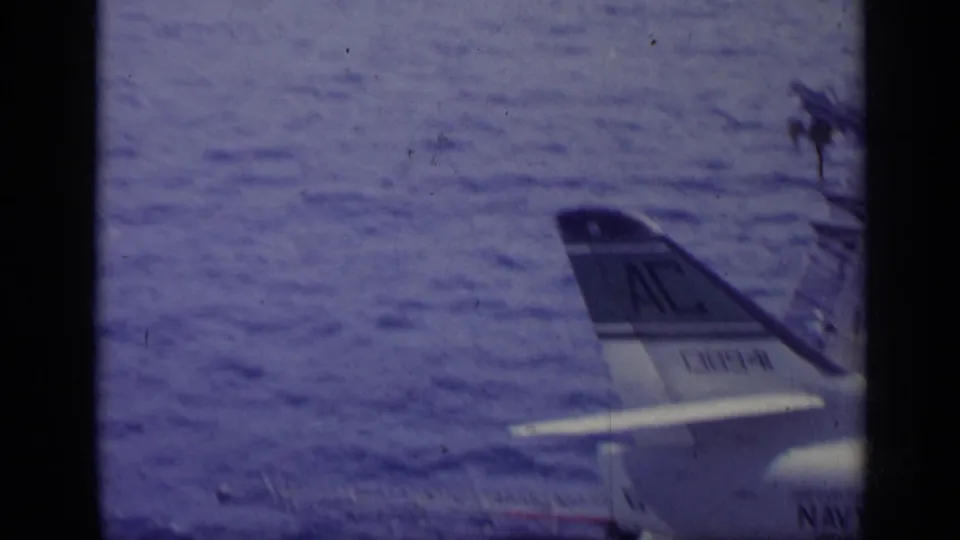 Aircraft tail on what appears to be a carrier deck, with blue-tinted water and a distant Italian shoreline beyond.