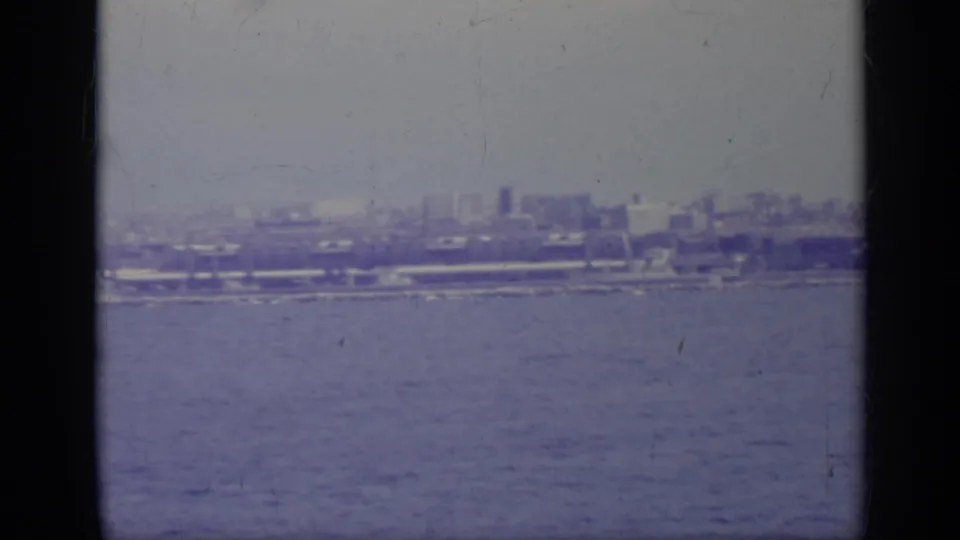 1962 8mm coastal view, likely in Italy per metadata, with water in the foreground and distant industrial buildings in haze.
