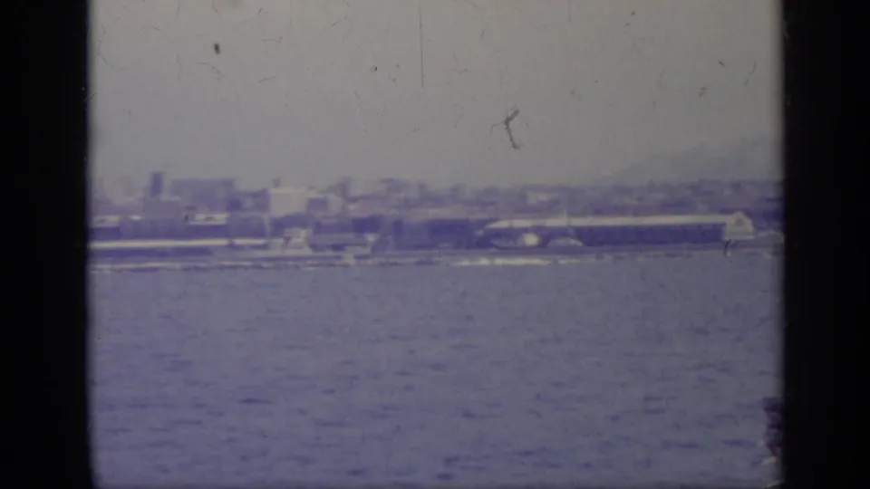 Faded 1962 8mm frame of a distant coastal harbor area, likely Italy per metadata, viewed across open water.
