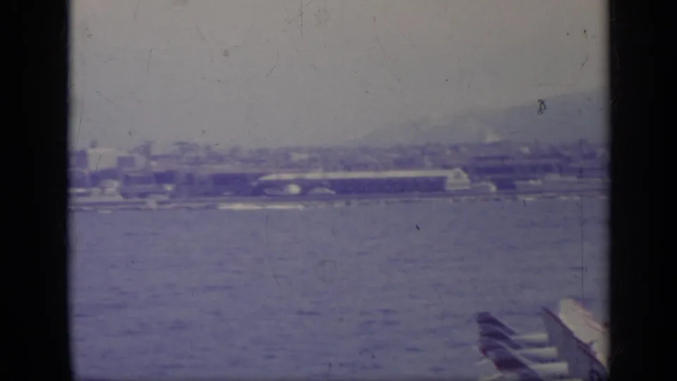 1962 8mm view across an Italian bay toward parked military aircraft, low waterfront buildings, and distant hills.