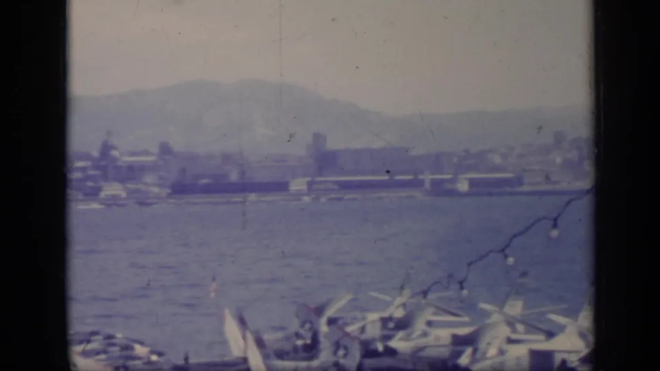1962 8mm frame from Italy showing parked aircraft near a bay, distant port buildings, hills, and a string of lights crossing the view.