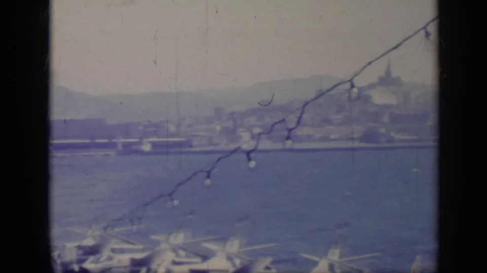 1962 Italian bay view with hanging lights in the foreground, aircraft below, and a hazy hillside port beyond.