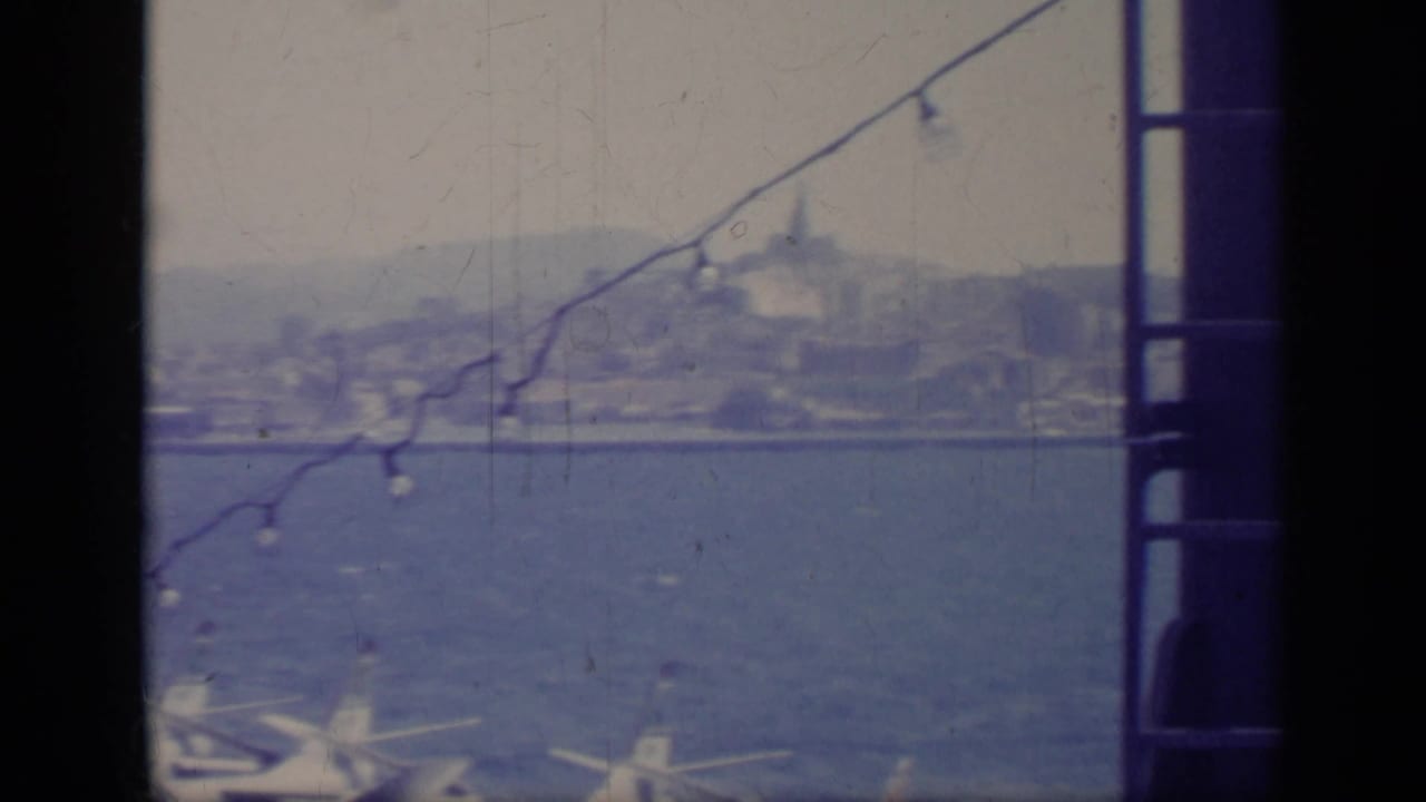 Distant shoreline and hills across blue water, seen through dark vessel framing and diagonal string lights.