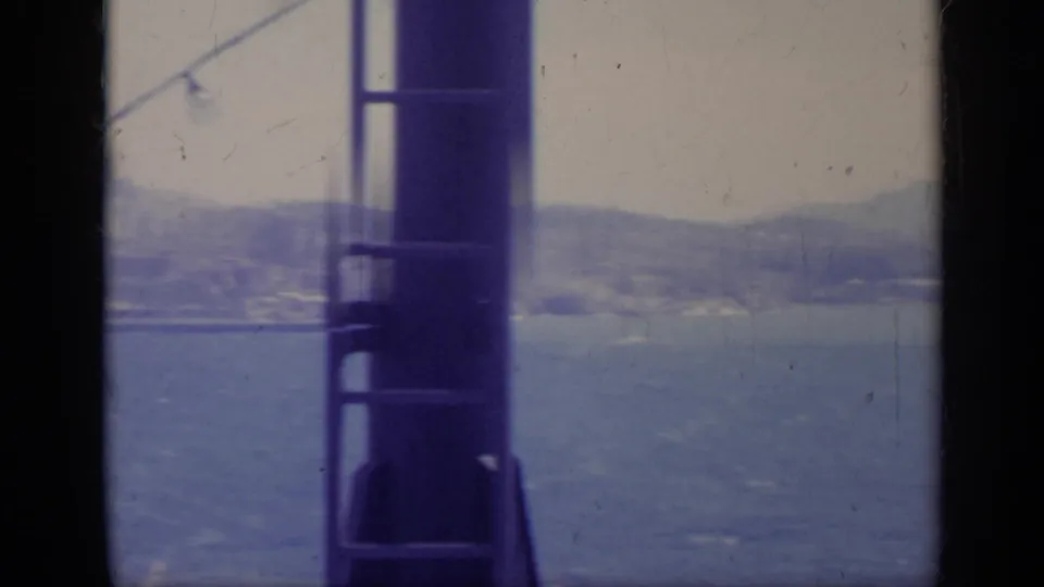1962 8mm Italy coastline across blue water, with mountains and shoreline partly obscured by dark vessel framing.