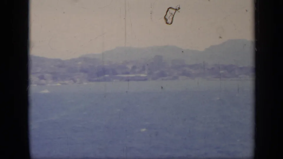 1962 8mm Italy home-movie view of a hazy shoreline and hills across blue water, framed by dark vertical film or window edges.