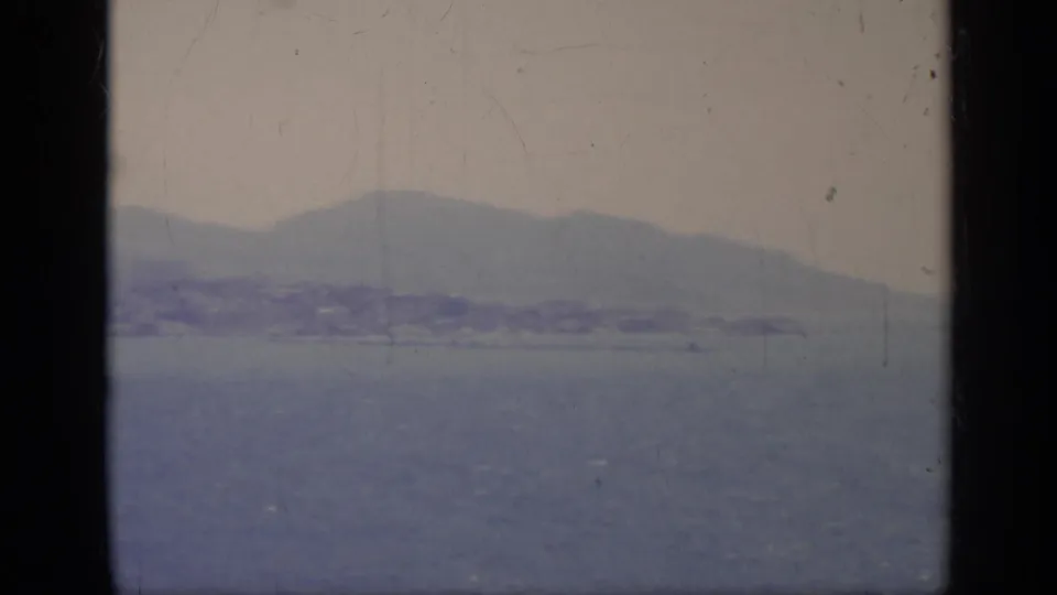 1962 8mm Italy coastal landscape with blue water, a distant mountain ridge, and shoreline viewed through dark foreground framing.