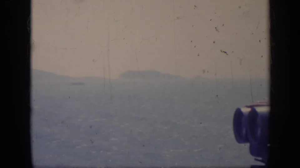 A hazy 1962 Italy seaside view with distant land across the water and binoculars partly visible at frame right.