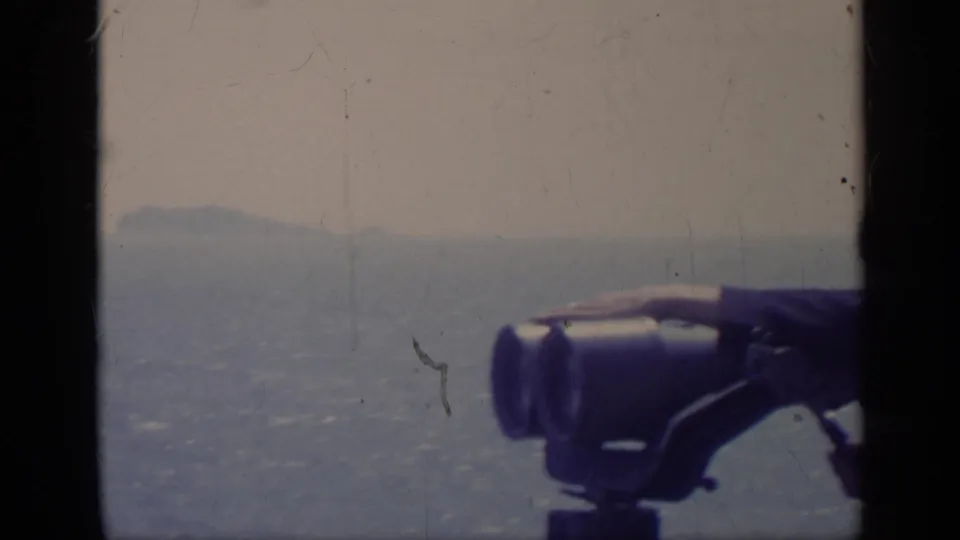 1962 Italy home-movie frame with a hand adjusting mounted binoculars against a soft coastal horizon.