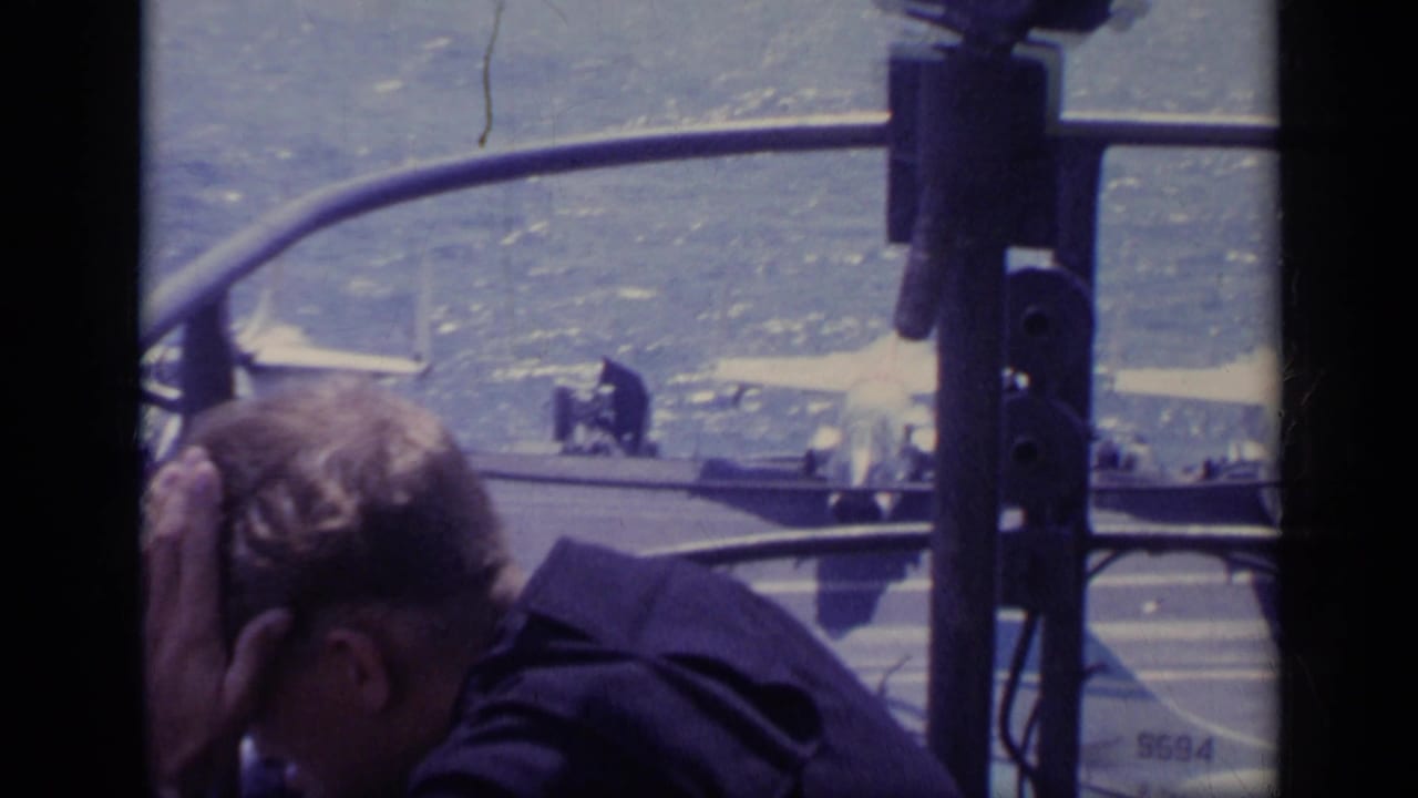 Man leaning in foreground while jet aircraft sit on a carrier deck beyond railings.