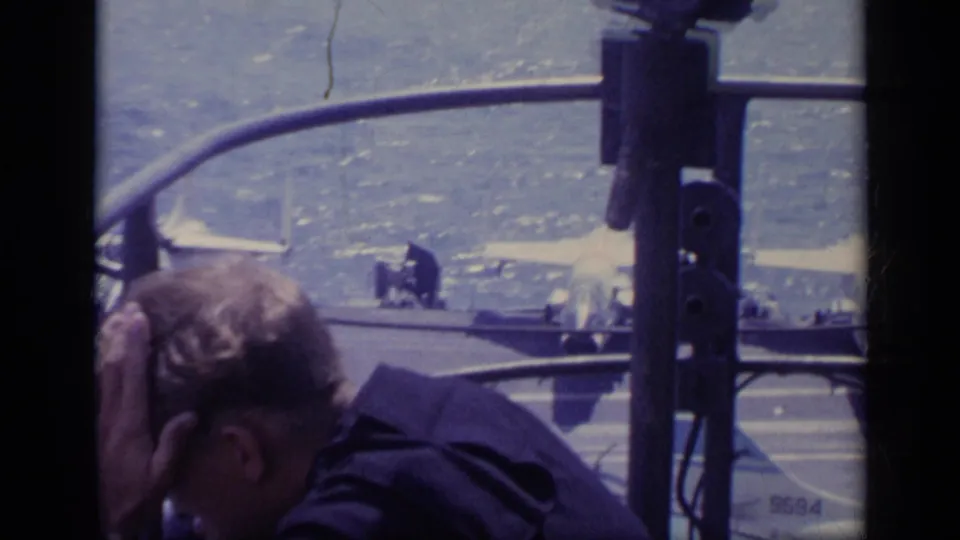 In Italy 1962 footage, a man leans forward behind railings as parked jet aircraft and ocean are seen on an aircraft carrier deck.